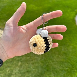 May include: A crocheted bee keychain with black and white stripes and white pom-poms for antennae. The bee is made of a soft, fuzzy material and has a black button eye.