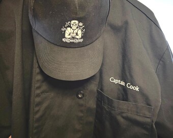 Puede incluir: Gorra de béisbol negra con un diseño de calavera y huesos cruzados y el texto "Captain Cook's Wood". También se ve una camisa negra con botones con el texto "Captain Cook" en el bolsillo del pecho.