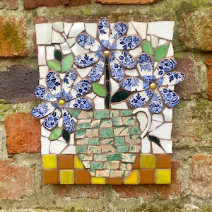 Könnte beinhalten: Ein Mosaikkunstwerk, das eine Vase mit blau-weißen Blumenmustern zeigt. Die Blumen sind aus zerbrochenem Porzellan gefertigt, und die Vase ist grün und beige. Das Kunstwerk ist an einer Backsteinmauer angebracht.