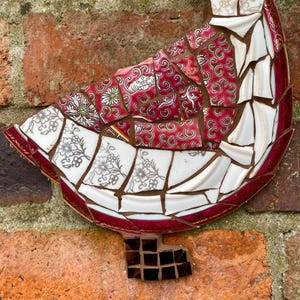 Pu&ograve; includere: Una scultura di uccello a mosaico realizzata con pezzi di porcellana rotti, con un motivo floreale rosso e bianco. L'uccello &egrave; montato su un muro di mattoni.