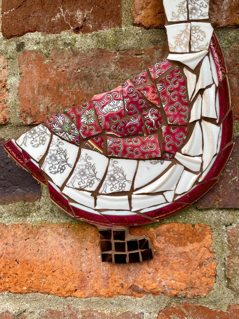 Pu&ograve; includere: Una scultura di uccello a mosaico realizzata con pezzi di porcellana rotti, con un motivo floreale rosso e bianco. L'uccello &egrave; montato su un muro di mattoni.
