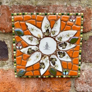 Puede incluir: Mosaico cuadrado con diseño de girasol. El mosaico utiliza azulejos naranjas y trozos de un plato con estampado floral. El centro del girasol es un plato con un escudo. Piezas de vidrio verdes y azules añaden acentos.
