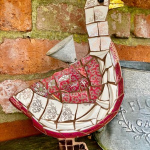 Pu&ograve; includere: Una scultura di oca a mosaico realizzata con pezzi di porcellana e ceramica rotti. L'oca &egrave; rosa, bianca e dorata con un occhio nero e un becco giallo. L'oca &egrave; in piedi su una superficie di legno.