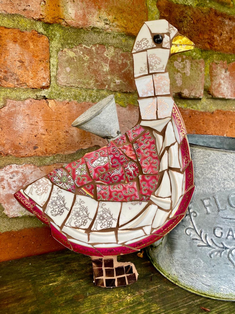 Pu&ograve; includere: Una scultura di oca a mosaico realizzata con pezzi di porcellana e ceramica rotti. L'oca &egrave; rosa, bianca e dorata con un occhio nero e un becco giallo. L'oca &egrave; in piedi su una superficie di legno.