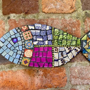Könnte beinhalten: Eine Mosaik-Fischskulptur mit farbenfrohem Design. Der Fisch besteht aus verschiedenen Fliesen in Blau-, Grün-, Lila- und Weißtönen mit einem dekorativen Auge. Das Mosaik ist an einer Ziegelmauer angebracht und zeigt die Textur und die Details des Kunstwerks.