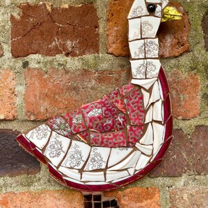 Pu&ograve; includere: Un'oca a mosaico realizzata con pezzi di porcellana e ceramica rotti. L'oca &egrave; bianca con accenti rossi e neri. L'oca &egrave; montata su un muro di mattoni.