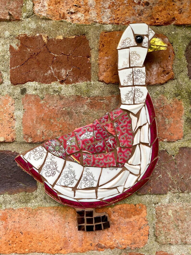Pu&ograve; includere: Un'oca a mosaico realizzata con pezzi di porcellana e ceramica rotti. L'oca &egrave; bianca con accenti rossi e neri. L'oca &egrave; montata su un muro di mattoni.