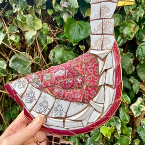 Pu&ograve; includere: Una scultura di oca a mosaico realizzata con pezzi di porcellana rotti. L'oca &egrave; bianca con un bordo rosa e dorato. L'oca &egrave; in piedi su una base marrone.