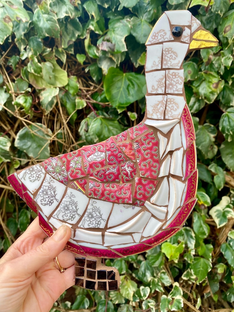 Pu&ograve; includere: Una scultura di oca a mosaico realizzata con pezzi di porcellana rotti. L'oca &egrave; bianca con un bordo rosa e dorato. L'oca &egrave; in piedi su una base marrone.
