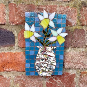 May include: A rectangular mosaic wall art piece featuring three white and yellow flowers in a vase. The background is composed of small blue tiles, while the vase is crafted from fragments of white ceramic with a floral pattern and green stems.
