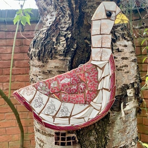 Pu&ograve; includere: Un'oca a mosaico realizzata con pezzi di porcellana e piastrelle di ceramica. L'oca &egrave; rossa, bianca e dorata con un occhio nero e un becco giallo. L'oca &egrave; attaccata a un tronco d'albero.