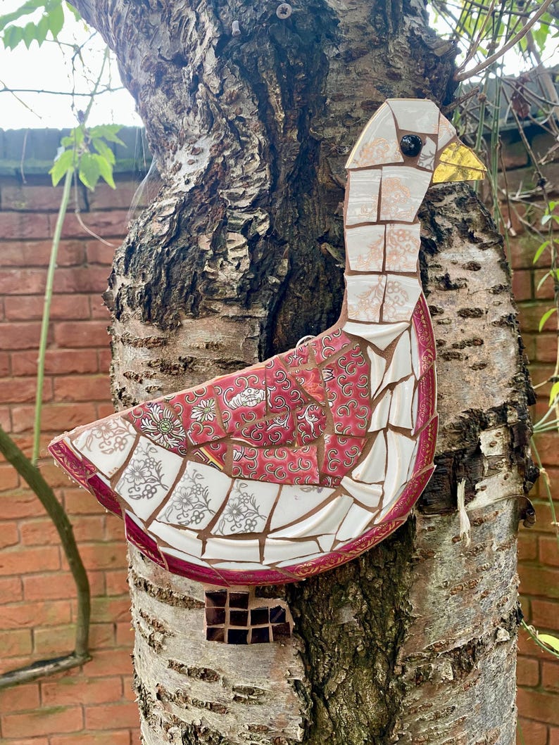 Pu&ograve; includere: Un'oca a mosaico realizzata con pezzi di porcellana e piastrelle di ceramica. L'oca &egrave; rossa, bianca e dorata con un occhio nero e un becco giallo. L'oca &egrave; attaccata a un tronco d'albero.