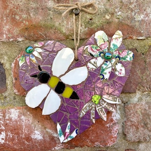 May include: A heart-shaped mosaic wall hanging featuring a bee and floral accents. The purple mosaic heart has a bee with white wings and a yellow and black body. Floral elements with green and blue accents are also included.