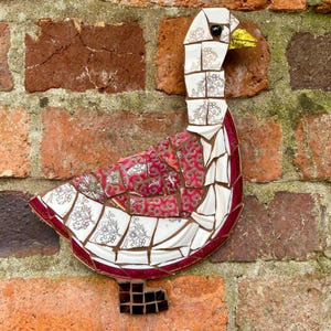 Pu&ograve; includere: Un'oca a mosaico realizzata con pezzi di piastrelle di ceramica rotte. L'oca &egrave; bianca con accenti rossi e dorati. &Egrave; montata su un muro di mattoni.