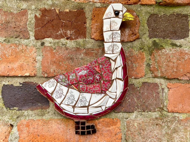 Pu&ograve; includere: Un'oca a mosaico realizzata con pezzi di piastrelle di ceramica rotte. L'oca &egrave; bianca con accenti rossi e dorati. &Egrave; montata su un muro di mattoni.