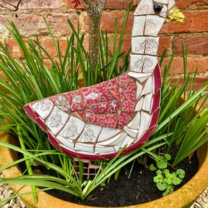 Pu&ograve; includere: Una scultura di oca a mosaico realizzata con porcellana rotta e piastrelle di ceramica. L'oca &egrave; bianca con accenti rosa e dorati e si trova in un vaso di terracotta pieno di piante verdi.