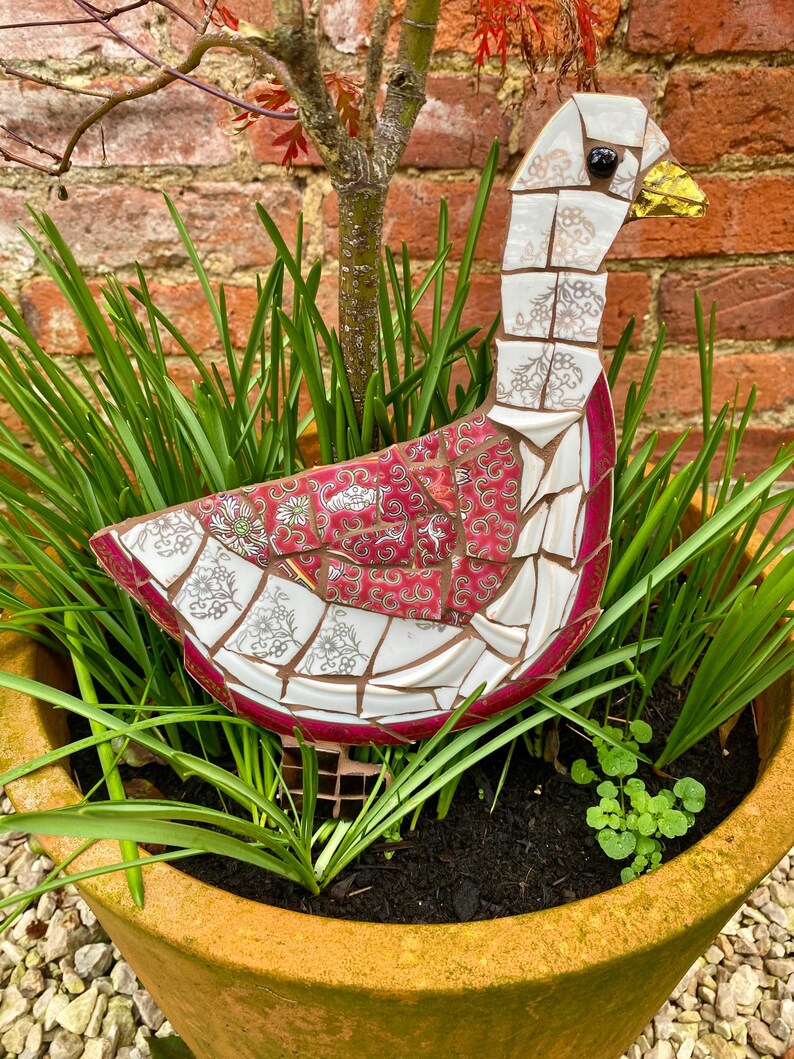 Pu&ograve; includere: Una scultura di oca a mosaico realizzata con porcellana rotta e piastrelle di ceramica. L'oca &egrave; bianca con accenti rosa e dorati e si trova in un vaso di terracotta pieno di piante verdi.