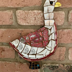 Pu&ograve; includere: Un'oca a mosaico realizzata con piatti di porcellana rotti. L'oca &egrave; bianca con accenti rossi e dorati. L'oca &egrave; montata su un muro di mattoni.