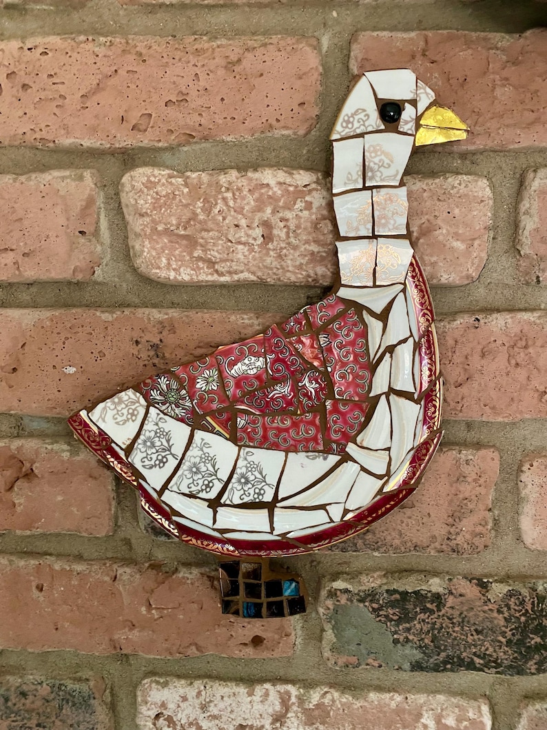 Pu&ograve; includere: Un'oca a mosaico realizzata con piatti di porcellana rotti. L'oca &egrave; bianca con accenti rossi e dorati. L'oca &egrave; montata su un muro di mattoni.