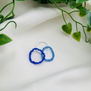 May include: Three beaded rings, one white and two blue, on a white surface.