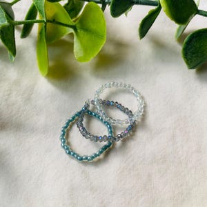 May include: Three beaded rings in shades of blue and clear. The rings are made of small, round beads strung together. The rings are arranged on a white surface with green leaves in the background.