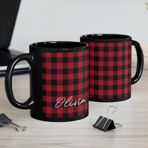 May include: Two black coffee mugs with a red and black buffalo plaid pattern. One mug has the name "Olivia" in a white script font. The mugs are on a light wood surface.