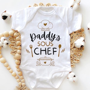 May include: White baby onesie with gold lettering that says "Daddy's Sous Chef" with a chef hat, fork, spoon, and pot graphic.