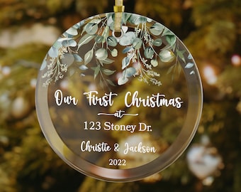 May include: Clear glass Christmas ornament with gold trim and a gold ribbon for hanging. The ornament features the text "Our First Christmas at 123 Stoney Dr. Christie & Jackson 2022" and is decorated with green and white floral accents.
