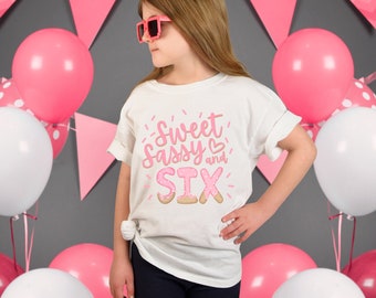 May include: A white t-shirt with the text "Sweet Sassy and SIX" in pink and light brown. The shirt is worn by a person with pink sunglasses, surrounded by pink and white balloons and pink triangle banners.