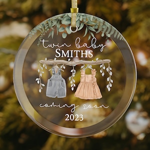 May include: A clear glass ornament with a green leafy design around the edge. The ornament features a branch with two baby clothes hanging from it, one blue and one pink. The text "twin baby SMITHS coming soon 2023" is printed on the ornament.