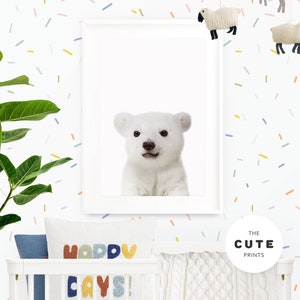 Puede incluir: Una impresi&oacute;n enmarcada blanca de un cachorro de oso polar con un fondo blanco. La impresi&oacute;n est&aacute; colgada en una pared blanca con confeti de colores. La pared tambi&eacute;n tiene una cuna blanca con una manta de punto blanco y una almohada con el texto "Happy Days!" en letras de colores.