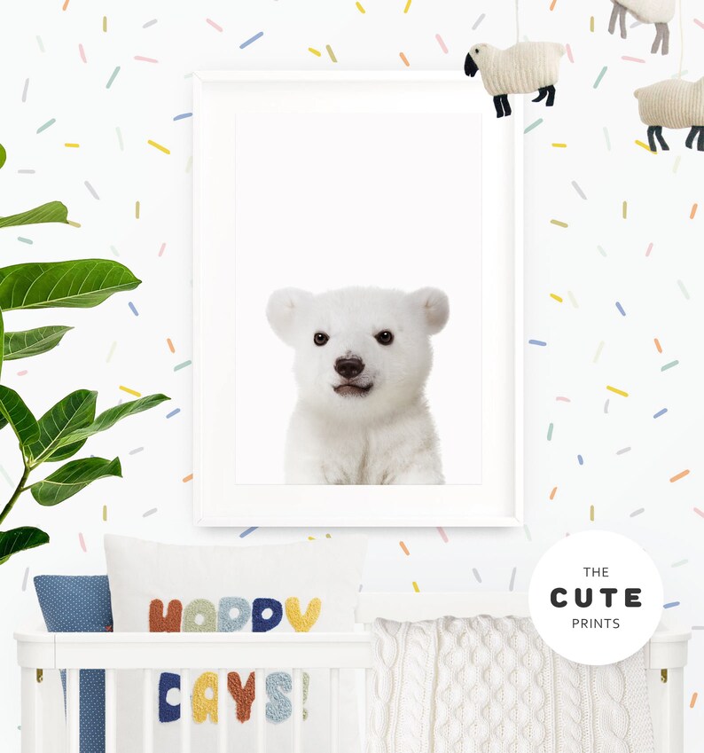 Puede incluir: Una impresi&oacute;n enmarcada blanca de un cachorro de oso polar con un fondo blanco. La impresi&oacute;n est&aacute; colgada en una pared blanca con confeti de colores. La pared tambi&eacute;n tiene una cuna blanca con una manta de punto blanco y una almohada con el texto "Happy Days!" en letras de colores.
