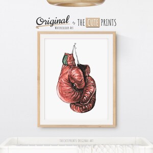 May include: Watercolor art print of red boxing gloves hanging against a white background. The gloves are detailed with shading and texture, and are suspended by a string. The print is framed in a light wood frame. The text "Original by THE CUTE PRINTS" is at the top.