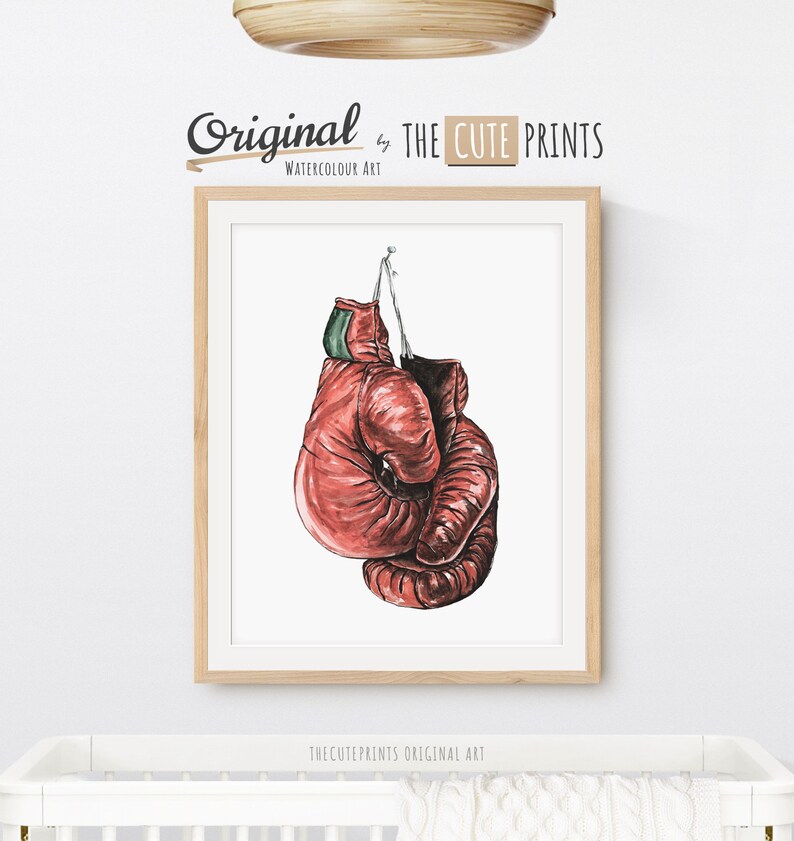 May include: Watercolor art print of red boxing gloves hanging against a white background. The gloves are detailed with shading and texture, and are suspended by a string. The print is framed in a light wood frame. The text "Original by THE CUTE PRINTS" is at the top.