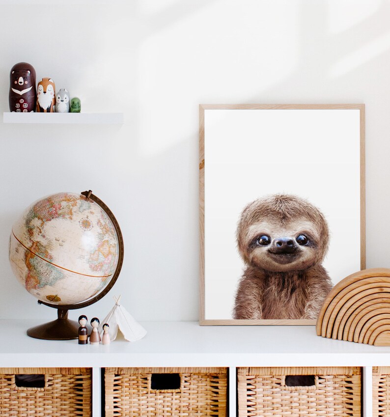 May include: A framed print of a sloth with a white background. The sloth is looking directly at the camera and has a happy expression. The frame is made of wood and has a light brown finish.