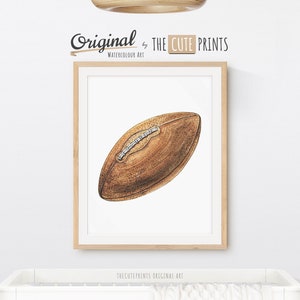 May include: Watercolor art print of a brown football with white laces, framed in a light wood frame. The print is labeled "Original by THE CUTE PRINTS WATERCOLOUR ART". The print is displayed above a white crib.