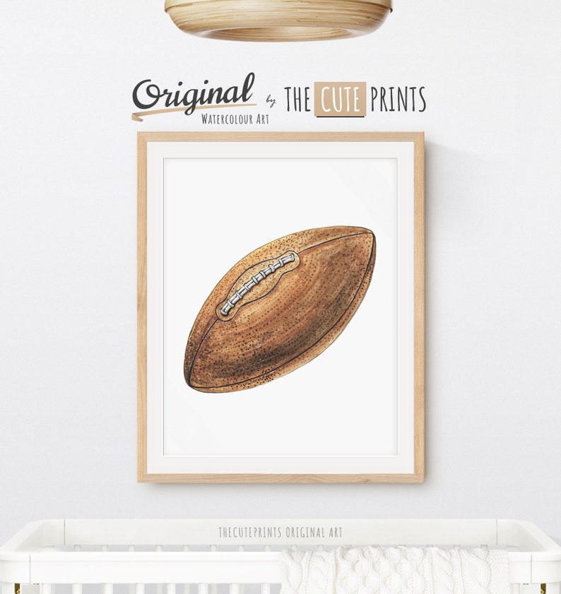 May include: Watercolor art print of a brown football with white laces, framed in a light wood frame. The print is labeled "Original by THE CUTE PRINTS WATERCOLOUR ART". The print is displayed above a white crib.
