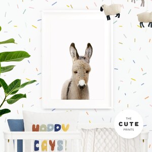 May include: A white framed print of a cute donkey with a white background. The print is hanging on a white wall with a colourful confetti pattern. The print is in a nursery setting with a white cot and a pillow with the text "Happy Days!" on it.