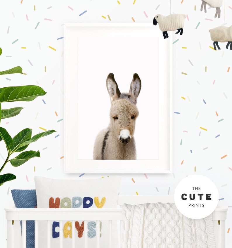 May include: A white framed print of a cute donkey with a white background. The print is hanging on a white wall with a colourful confetti pattern. The print is in a nursery setting with a white cot and a pillow with the text "Happy Days!" on it.