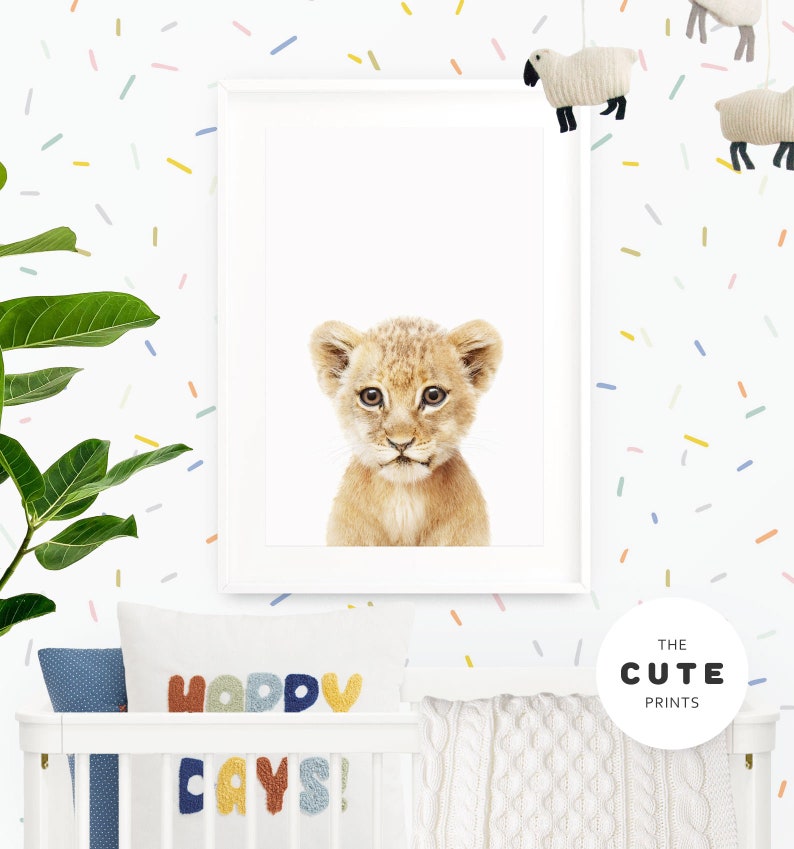 Peut inclure: Une impression encadr&eacute;e d'un adorable lionceau sur fond blanc. L'impression est accroch&eacute;e &agrave; un mur blanc avec des confettis multicolores. Le mur a &eacute;galement un lit b&eacute;b&eacute; avec une couverture en tricot blanc et un oreiller avec les mots "Happy Days!" en lettres color&eacute;es.