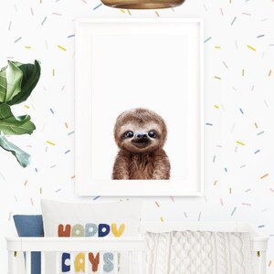 May include: A framed print of a smiling sloth with a white background. The print is hanging on a white wall with a colorful confetti pattern. Below the print is a white crib with a blue and white patterned pillow and a colorful pillow with the text "Happy Days!"
