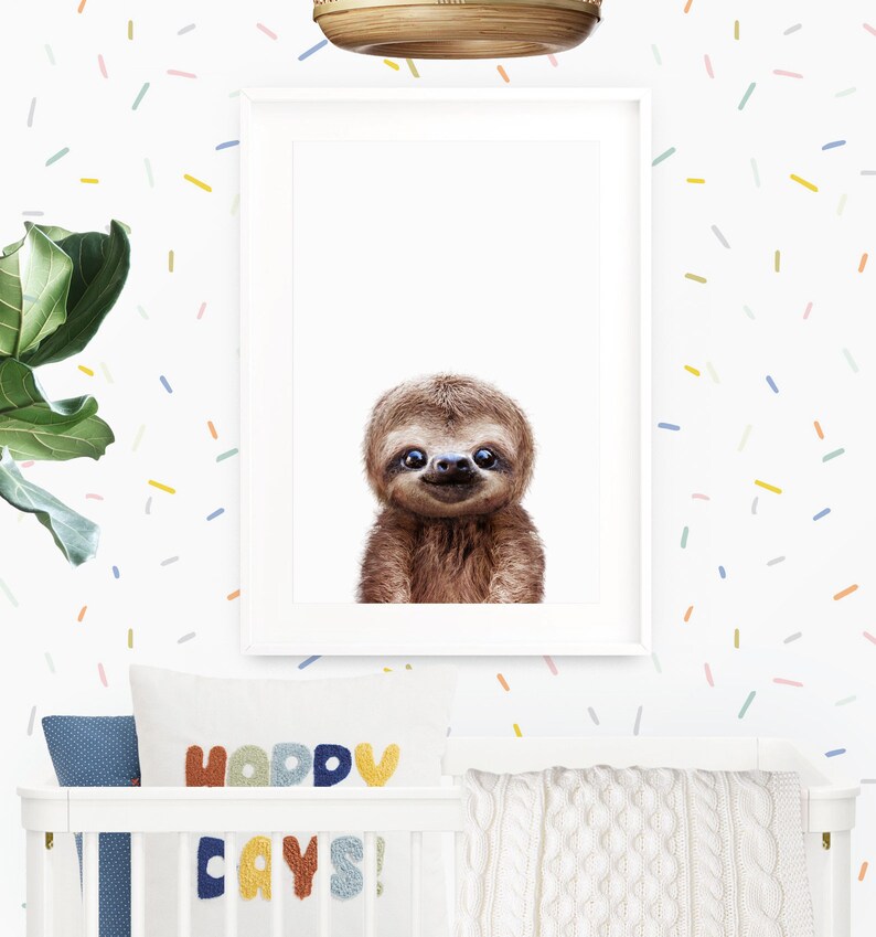 May include: A framed print of a smiling sloth with a white background. The print is hanging on a white wall with a colorful confetti pattern. Below the print is a white crib with a blue and white patterned pillow and a colorful pillow with the text "Happy Days!"