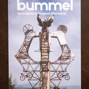 Könnte beinhalten: Magazin-Cover mit dem Titel "bummel" in großen weißen Buchstaben. Das Cover zeigt eine große, abstrakte Metallskulptur mit zwei kleinen Fahrrädern. Der Text "An irregular zine about bike travel" ist ebenfalls sichtbar.