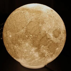 May include: A spherical, textured lamp resembling the moon. The lamp has a warm, yellow glow and sits on a white base. The surface of the lamp is detailed with craters and other lunar features. A black power cord extends from the base.