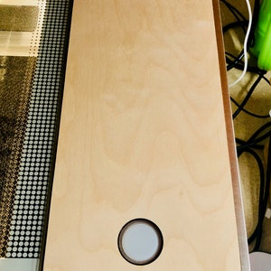 May include: A light-colored wooden surface with a circular inset, set within a metal frame. The wood grain is visible. The surface is part of a larger machine, with a patterned section visible on the left.