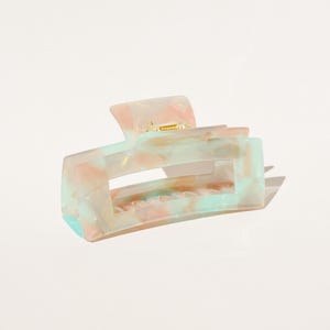 Peut inclure: Une pince à cheveux rectangulaire aux couleurs pastel. La pince présente un motif marbré avec des nuances de rose, de vert menthe et de crème. La pince a une charnière et des dents en métal doré pour saisir les cheveux.