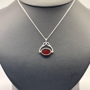 Può includere: Una collana con pendente in argento con una gemma ovale rosso scuro. La gemma è incastonata in una cornice d'argento decorativa e pende da una delicata catena d'argento. La collana è esposta su un manichino nero.