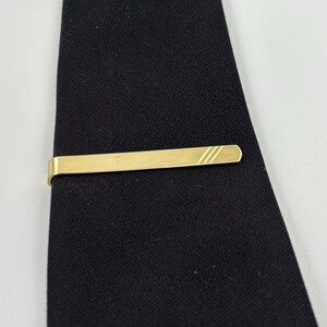 May include: A gold-coloured tie clip with three diagonal lines on one end, resting on a black tie. The clip is rectangular with rounded edges. The tie has a textured appearance.