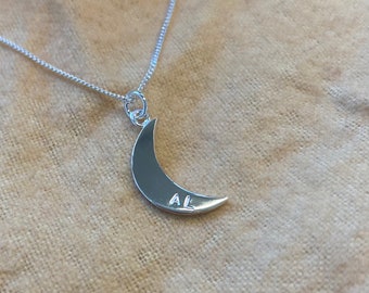 May include: A silver necklace with a crescent moon pendant. The pendant has the initials "AL" engraved on it.