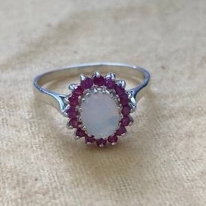 Op de afbeelding: Een zilveren ring met een witte ovale opaal steen omringd door een halo van kleine roze edelstenen.